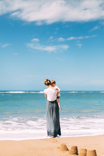 Γυναίκα κρατά το παιδί της στην αγκαλιά της στην παραλία δίπλα στη θάλασσα.