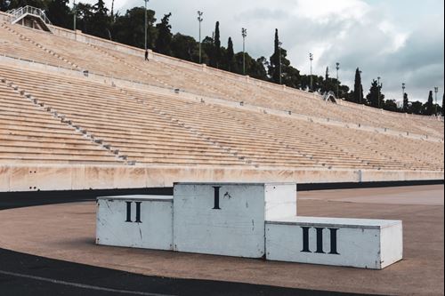 Στη φωτoγραφία φαίνεται η μία πλευρά των κερκίδων του Καλλιμάρμαρου σταδίου. Σε πρώτο πλάνο το τριπλό βάθρο των νικητών, όπου φαίνονται, από αριστερά προς τα δεξιά, οι αριθμοί 2, 1, 3 στα λατινικά.
